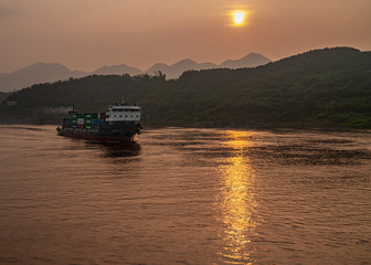 Chongqing, China - May 8, 2010: Evening light on Yangtze River. Container ship sails on while golden sunset reflected by brown water in front of shoreline with sun above forested hills.