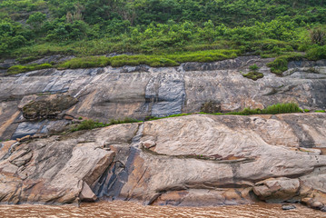 Chongqing, China - May 8, 2010: Evening light on Yangtze River. Closeup of flowing water blackening parts of brown-gray rock slates of shoreline behind brown water. Green vegetation on top.