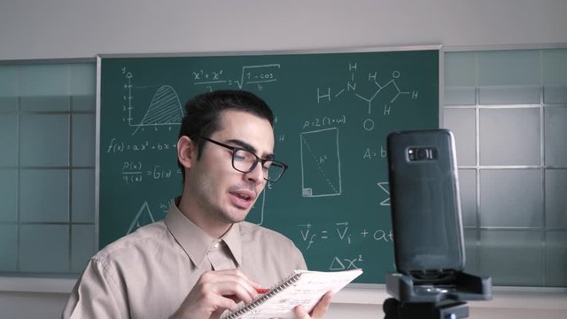 A male Caucasian online educator , with a chalk board behind him , teaching mathematics to his students via video call on  his smartphone mobile device