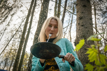 Young blonde woman is sitting in a summer forest on a fallen tree, holding a pan with fried eggs and sausages and eating. Concept of tourism and travel.
