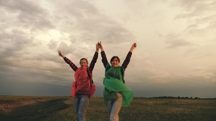 Fototapeta na wymiar strong and healthy girls travelers travel in colorful raincoats, climb the mountain, hold hands, help each other. Free women tourists with backpacks on rise to top of hill, rejoice in victory and jump