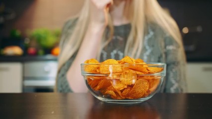 Glad woman eating potato chips. Beautiful young female enjoying potato chips and looking at camera while sitting in stylish kitchen.
