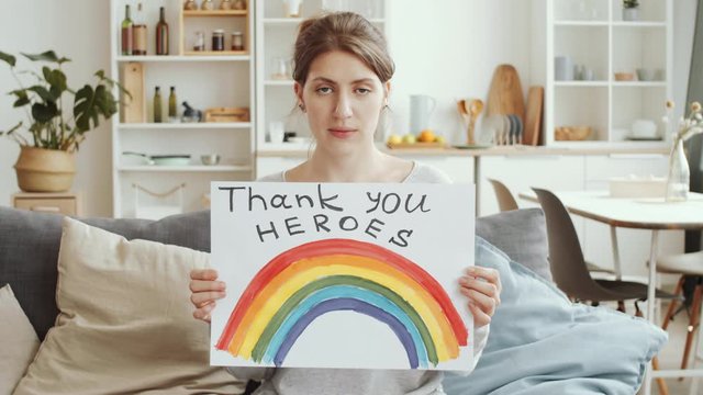 Young Caucasian woman showing handmade poster with rainbow and «NHS thank you heroes» inscription and looking at camera while sitting on couch at home during coronavirus quarantine