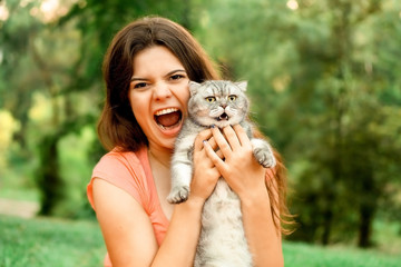 Beautiful long-haired brunette girl in a park sitting with a cat. The girl growls at the cat and bites. Photoshoot with a pet cat on the street..