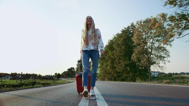 Cheerful woman with suitcase walking on road. From below shot of pretty young female smiling and pulling red suitcase while walking on asphalt road during trip through countryside.