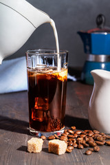 Cream is poured into a glass with iced coffee, on wooden table are coffee beans and sugar.