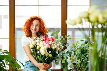 Young florist woman with red curly hair holding a freshly made blooming bouquet of flowers with ...