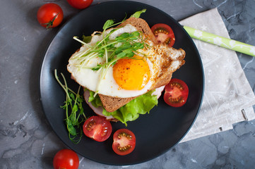 Sandwich with fried egg, meat, tomatoes, peas sprouts  and salad. Breakfast on a black plate. Top view
