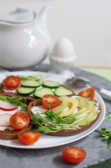 Vegetarian sandwiches with cream cheese, cucumber, avocado, garden radish, tomatoes and peas sprouts on white plate. Egg mist on white stand. Breakfast with vegetarian food.