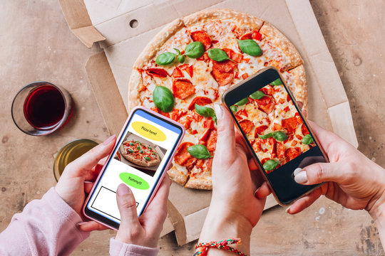 Female friends using smartphones to take photos of their pizza. Pepperoni Pizza in box with basil on rustic background. Italian food delivery, call or order online on mobile.