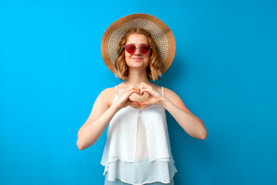 summer concept. girl in a sun hat and glasses shows a heart with her hands on a blue isolated background, a woman in summer clothes makes a gesture of love on vacation - Powered by Adobe