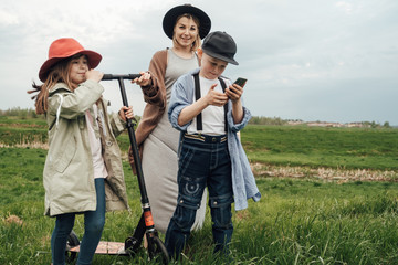 Happy family, mother, daughter and son in organic clothes, walk in the field and ride a scooter and watch video on the phone. The concept of a healthy lifestyle, happiness and joy