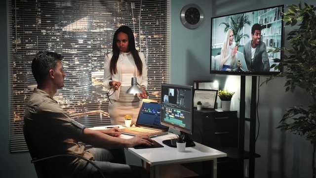 African-american chief director showing unsatisfied emotions to video editor's work. There is a man with green screen tablet in video watched on TV screen.