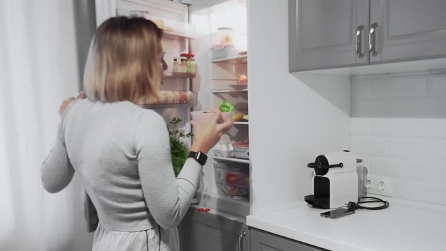 Woman opens refrigerator door in kitchen at home and takes salad