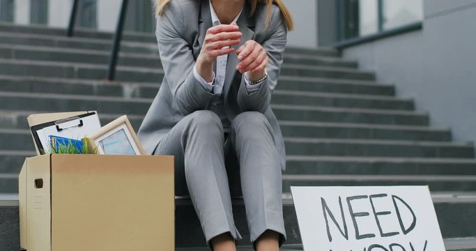 Portrait of Caucasian young workless woman siting on stairs at street with carton board Need Work and box with stuff. Female fired office worker in depression. Unemployment concept.
