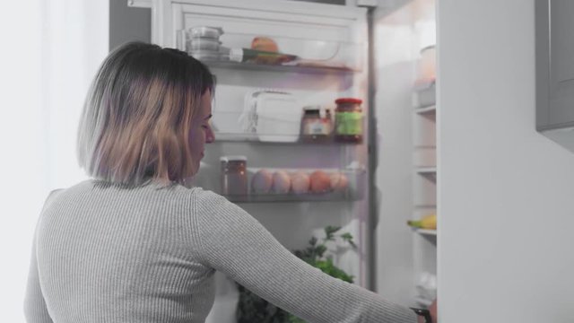 CLose up of Woman with headache or hangover opens refrigerator door in kitchen at home and takes bottle of water