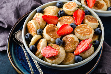 Trendy food - pancake cereal. Mini cereal pancakes with strawberries and blueberries in boul on blue cement background, top view. Horizontal format