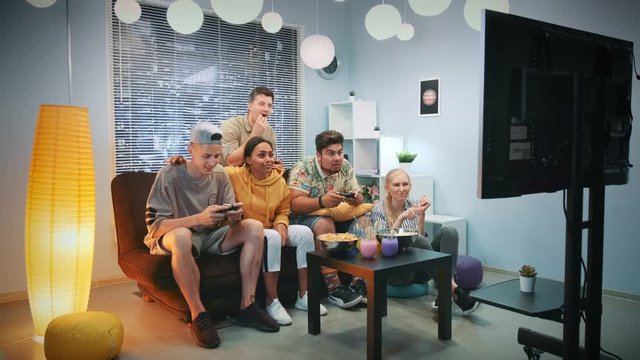Two gamers playing in console game among company of friends which cheering up them. They sit on sofa at nice home, eating snacks and having fun.
