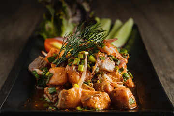 healthy food background of Thai style spicy raw salmon salad in black dish served with lettuce tomato radish and sliced cucumber, selective focused