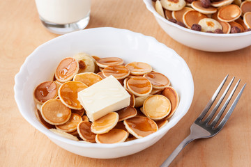 Trendy food during coronavirus quarantine - pancake cereal. Mini pancakes in white bowl with butter and honey, glass of milk and fork on wooden table. Breakfast for kids, tiny pancakes.