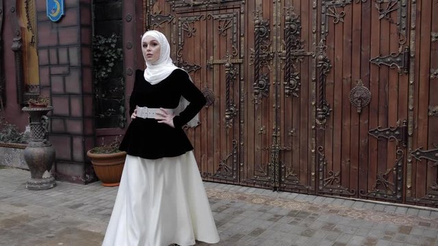 slim woman in elegant Arabian suit with white hijab walks away from wooden gate along restaurant yard slow motion