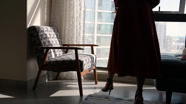 Woman with long brown dress walking in living room to chair next to window reading book at home over morning sunlight and building background.