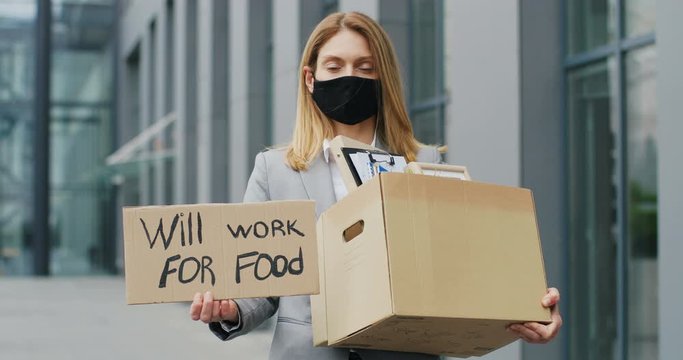 Portrait of Caucasian workless woman in mask standing outdoor and showing poster with words Will work for food. Fired female jobless and holding box with office stuff.