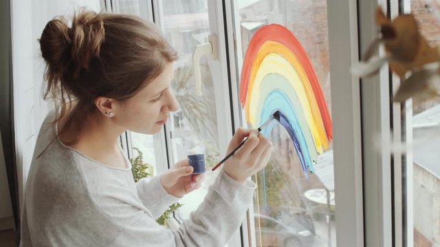 Side view of young creative woman drawing colorful rainbow with paint and brush on glass window while decorating home on quarantine