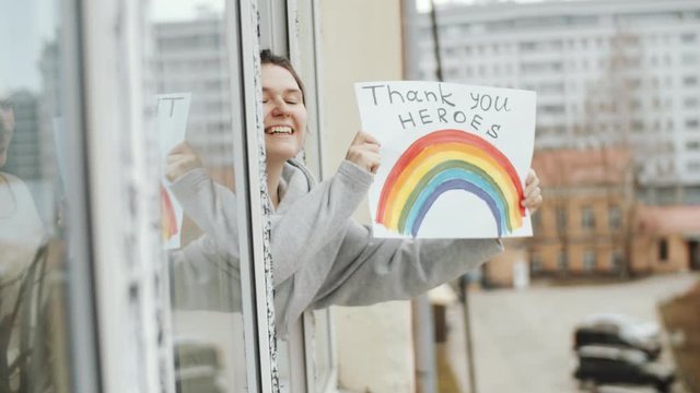 Young cheerful woman holding poster with inscription «NHS thank you heroes», looking out of the window, smiling and waving while expressing respect to healthcare workers during covid-19 pandemic