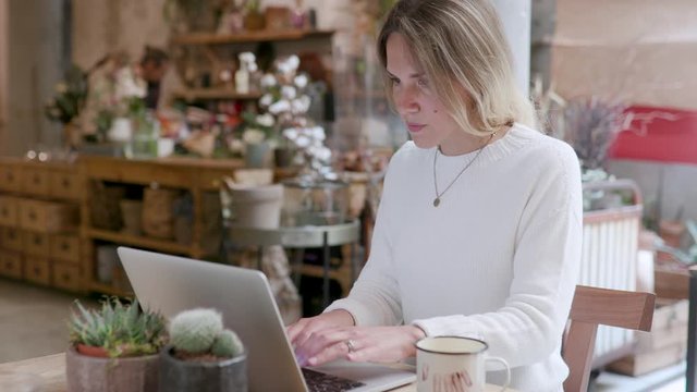 Young girl in remote working from a cafe bar, restaurant and flower shop. The woman is typing on the keyboard and scrolling on the trackpad on her laptop computer. Seated at the table, slow motion.