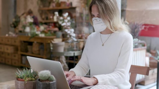 A woman with a protective mask is typing and scrolling on her laptop computer. Young girl in remote working from a cafe bar, restaurant and flower shop. Seated at the table, slow motion.