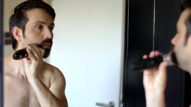 young man without clothes comes in the bathroom to the mirror and shaves his beard with a trimmer