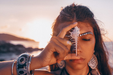 beautiful young tribal style woman holding sea shell at her face with eyes closed outdoors at sunset