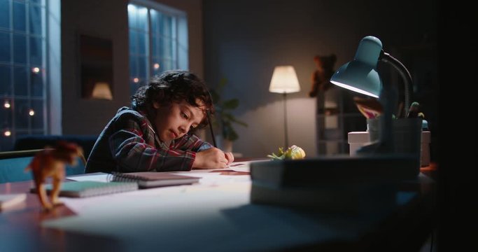 Funny little asian kid drawing at home. Boy with curly hair drawing with pencils on paper in the evening, learning art, having fun at home - hobby concept 4k footage