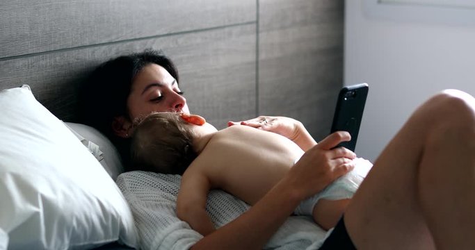 Mother using smartphone with baby sleeping on mom chest. Mom caressing napping infant while asleep