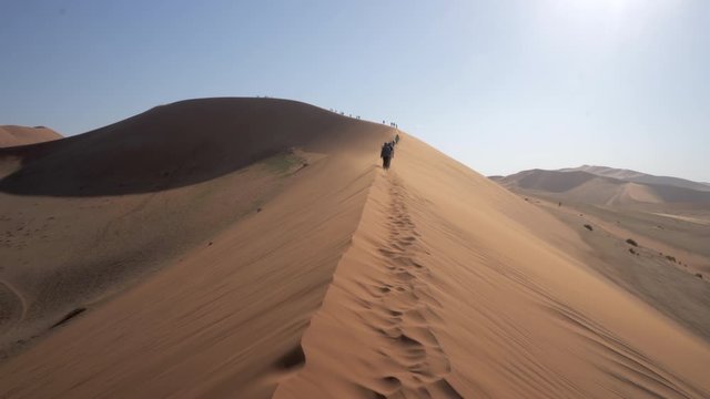 Tourists exploring desert while walking on sand dune against sky, people on arid landscape - Namib Desert, Namibia