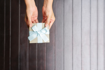 female hands holding a small gift wrapped with blue ribbon on wooden background