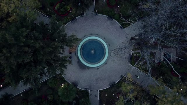 drone view of a fountain in the center of a park with trees and people walking