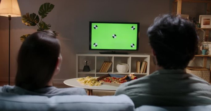 Couple spending time together in the evening, sitting on couch and watching tv with green chroma-key green screen. A template for your clips 4k footage
