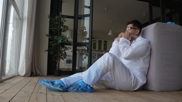 Tired man sitting on the floor in the apartment, in a protective suit, goggles and respirator