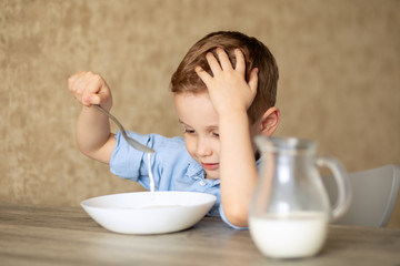 Adorable Caucasian baby girl eats porridge on her own. The boy does not like porridge. Playing with a spoon pours milk. Kids Healthy Food Concept. In room.