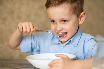 Adorable Caucasian child with brown eyes and red blond hair eats porridge himself. The boy bit his spoon with his teeth. He does not like milk porridge. The concept of eating healthy children.