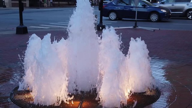 Lit running fountan in down town Lexington with traffic and buildings in the background at night.