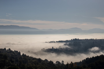 Obraz na płótnie Canvas Surreal view of of a little town in Umbria (Italy) almost completely hidden by fog with trees silhouettes in the foreground