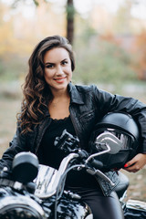 Plakat Young beautiful biker woman in a black leather jacket on a motorcycle with a helmet in his hands. The concept of speed and freedom. Soft selective focus.