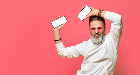Stylish positive grey-haired bearded adult man in white hoodie and sunglasses standing holding two smartphones in hands