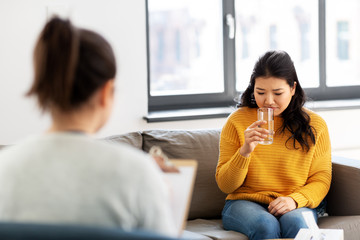 psychology and mental therapy concept - young asian woman patient drinking water and psychologist...