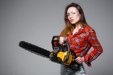 Naklejka premium Girl with a chainsaw on a gray background. Girl in a red shirt and construction safety glasses