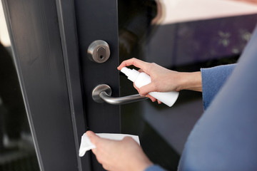 hygiene, health care and safety concept - close up of hands cleaning outdoor door handle surface with disinfectant spray and tissue