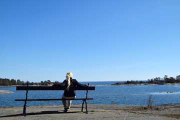 Blonde young woman, in her 30s, all alone, sitting on a bench, looking out to sea and the...